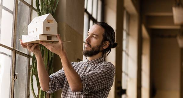 Mann in einem karierten Hemd steht vor einer Fensterfront und hält das Modell eines Hauses in die Sonne.